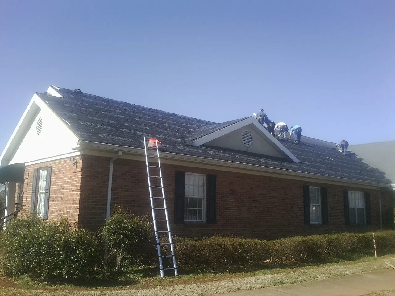 Crew tearing shingles off a brick ranch roof with a ladder set — Metal Roof Repair work by our crew in Rowley