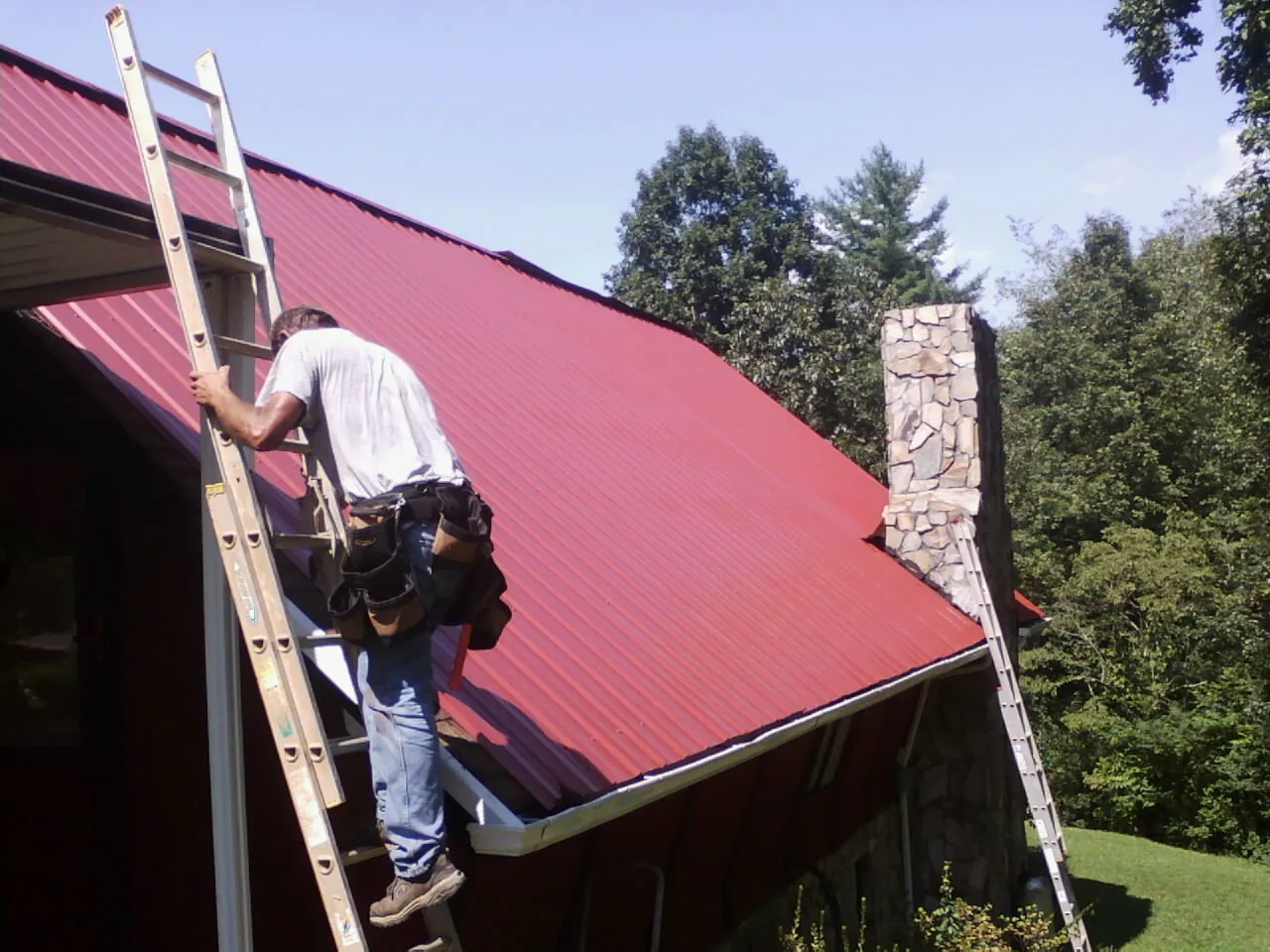 Roofer on a ladder beside a red metal roof and stone chimney — Soffit & Fascia Repair by Roofing Brothers in Rowley